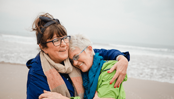 Deux amies s’enlacent sur la plage. Elles profitent à un âge avancé du rendement supplémentaire que leur assurance investissement leur a apporté.
