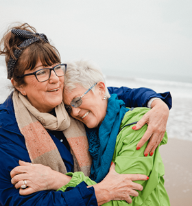 Twee vriendinnen omhelzen elkaar op het strand. Ze genieten op hun oude dag van het extra rendement dat hun beleggingsverzekering hen heeft opgebracht.