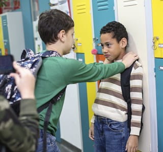Een jongen wordt gepest en tegen een kast geduwd, terwijl een andere jongen de situatie filmt met zijn smartphone.
