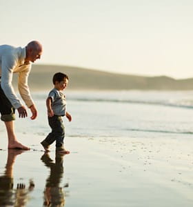 Un homme accompagné d’un enfant, se promenant au bord de plage et ayant l’esprit tranquille grâce à l’assurance voyage souscrite en Belgique.