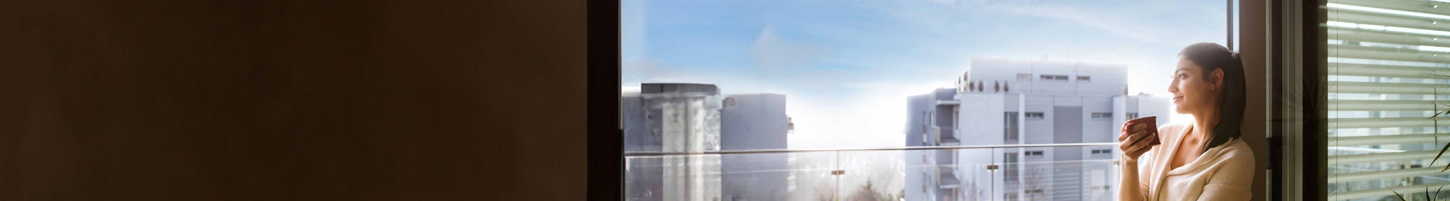 Grâce à son assurance immeuble, une femme profite sereinement d’un café, à la fenêtre de son appartement.