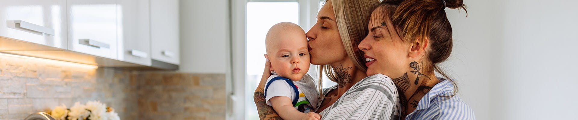 Een koppel vrouwen geniet van een knuffelmomentje met hun baby in de keuken. Ze hebben net een schuldsaldoverzekering afgesloten voor hun woning om elkaar te beschermen in geval van overlijden.